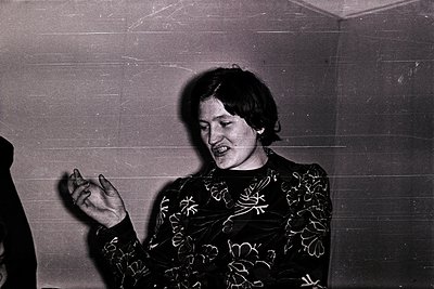 Black-and-white portrait of a person in mid-gesture, likely performing or speaking, wearing a patterned blouse with floral mo...