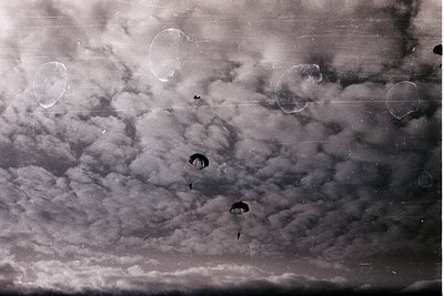 Vintage black-and-white aerial view of three parachutists descending through stormy clouds, likely mid-20th century. Circular...
