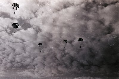 Black-and-white aerial view of paratroopers descending via silk parachutes during daylight. Dense cloud cover obscures ground...