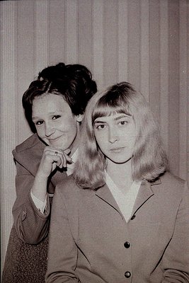 Two women in 1970s-style blouses pose indoors against a striped curtain. The woman on left wears a patterned jacket with a ro...