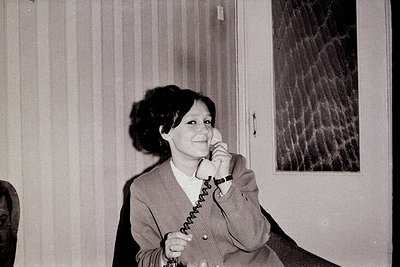 Vintage black-and-white photo of a woman in a 1970s-style blazer speaking on a rotary phone in an indoor office setting. Stri...