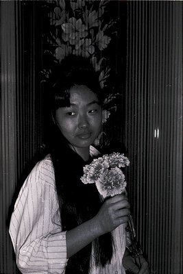 Black-and-white portrait of a woman in traditional East Asian attire, holding a bouquet of white flowers. Floral wallpaper wi...