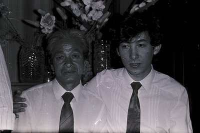 Two men pose indoors in formal attire, likely at a celebratory event. The elder wears a white shirt, striped tie, and a heads...