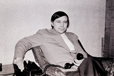 Portrait of a man in mid-20th-century formal attire—light-colored shirt, dark blazer, and trousers—seated in a plush armchair...
