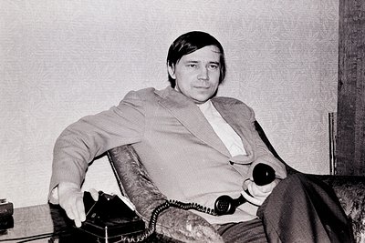 Black-and-white portrait of a man in a relaxed pose, seated in a leather armchair with a vintage rotary phone on his lap. His...