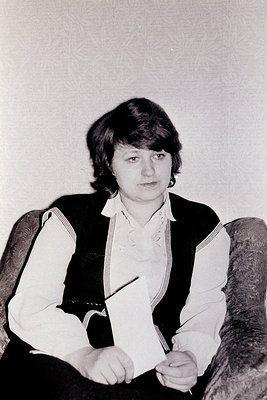 Portrait of an individual in a 1970s-style vest and blouse, seated indoors with a pen in hand. The patterned vest features di...