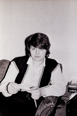 Portrait of a person in a vintage vest and striped suspenders, holding a folded paper or document. Background shows a vintage...