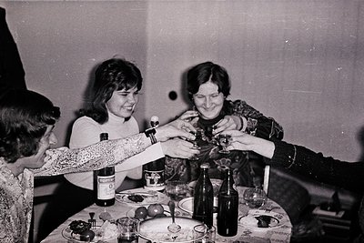 Two women in 1970s-era clothing share a toast at a table laden with bottles (including a visible "Bourbon" label) and glasses...