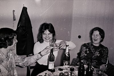 Vintage black-and-white indoor gathering featuring three women in 1970s-era attire. Central figure toasts with a bottle of *B...
