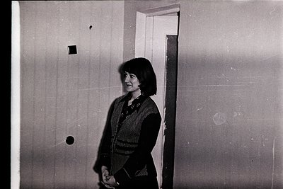 Black-and-white portrait of a woman in a striped sweater, standing indoors near a doorway with a light source behind her. Mid...