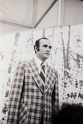 Mid-century formal portrait of a man in a checkered blazer and tie, standing indoors near a patterned wall. His expression is...