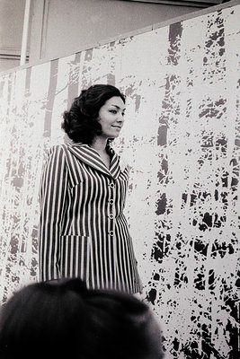Portrait of a woman in a striped blazer, posed indoors against a textured wall with abstract patterns. Her short, voluminous ...
