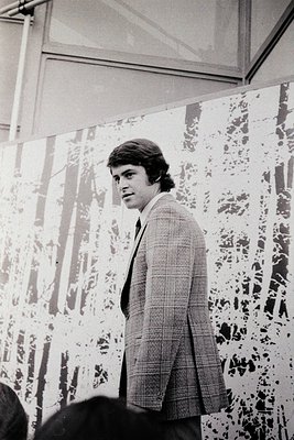 Young man in 1970s-style suit poses indoors near patterned wallpaper with geometric motifs. Mid-length hair and light-colored...
