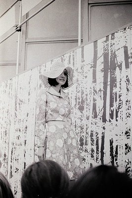 A woman in a floral dress and wide-brimmed hat poses against an abstract mural of tree-like splatters, likely from a 1960s–19...