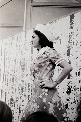 Mid-century fashion portrait: woman in 1950s–60s floral dress with white sailor-style hat, posing indoors with patterned wall...