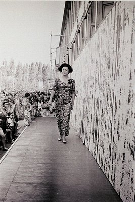 A model in a bold, floral-patterned 1960s-style jumpsuit strides down a runway flanked by spectators. The stark, industrial c...