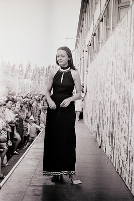 Black-and-white portrait of a woman in a floor-length halter dress with fringe trim, posing on a stage flanked by a crowd. Ar...