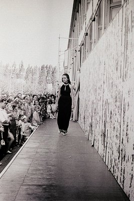 A woman in a black dress and high-necked top walks alone down a narrow, crowded corridor flanked by concrete walls. A large c...
