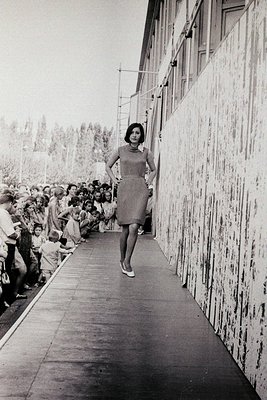 A woman in a sleek 1960s dress strides confidently down a narrow, crowded corridor flanked by concrete walls, holding a white...