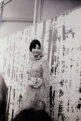 Portrait of a woman in a 1950s–60s evening gown with floral appliqué, posed indoors against a wall adorned with abstract, spl...