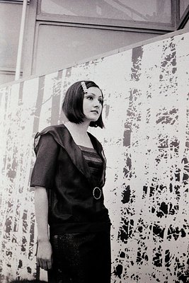 Portrait of a woman in a 1970s-style dress with a geometric leaf-patterned wall backdrop. Short hair, dark bob with side part...