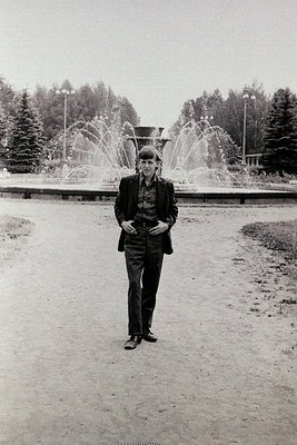 Black-and-white portrait of a man in a suit and hat, standing confidently in a landscaped park with a prominent fountain and ...