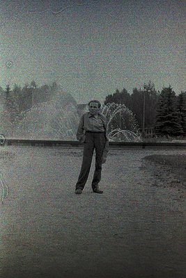 Black-and-white portrait of a man in mid-20th-century attire (wide-collar shirt, trousers) standing beside a cascading founta...
