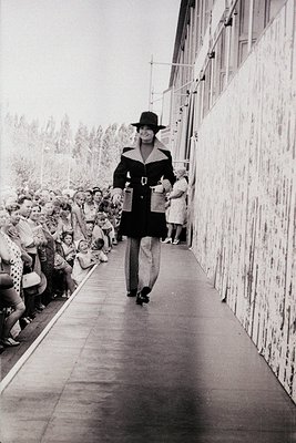 Fashion model strides down a runway flanked by spectators in a 1960s-70s runway show. Black-and-white print captures a sleek,...