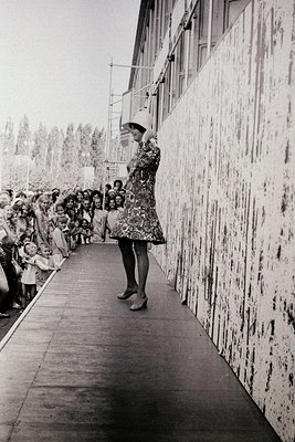 A woman in a floral dress and wide-brimmed hat poses mid-step on a concrete pathway beside a large, weathered concrete wall. ...