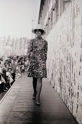 A model strides down a runway in a 1960s-style floral dress and wide-brimmed hat, flanked by a crowd. Industrial concrete wal...