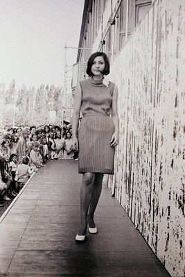 A model in a 1960s-style striped dress and tights strides down a runway flanked by a crowd. Industrial-style concrete walls a...