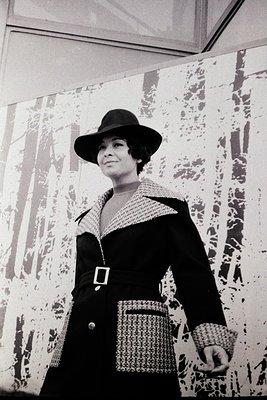 Elegant 1960s fashion portrait featuring a woman in a tailored black coat with a textured collar, wide-brim hat, and a beaded...