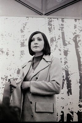 Portrait of a woman in a textured, high-collar coat, standing against a wall with abstract, splatter-paint-style artwork. Her...