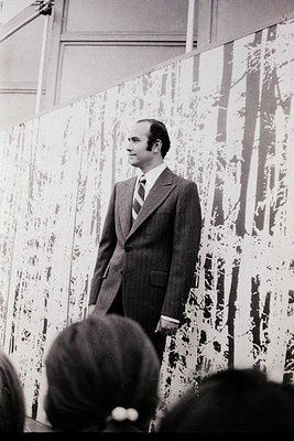 Man in a pinstripe suit and striped tie addresses an audience in a gallery setting, framed by abstract, textured wall art res...