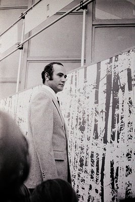 Man in light suit stands beside abstract splatter-paint artwork, likely 1960s–70s. Industrial gallery space with exposed beam...