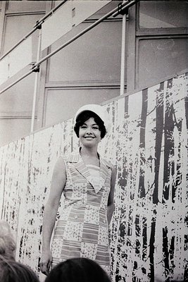 A woman in a 1960s-style dress with a fitted bodice and floral pattern stands indoors beside abstract art—vertical splatter p...