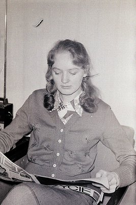 Mid-century woman reading a magazine, likely *Ladies' Home Journal* or similar, in a cozy indoor setting. She wears a buttone...