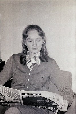 Young woman in 1970s-era blouse and cardigan reads *Bieta* magazine, likely from Eastern Europe. Plain interior suggests dome...