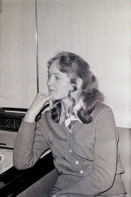 Portrait of a woman in a 1970s-style sweater and floral collar, seated indoors with a vintage radio or tape recorder beside h...