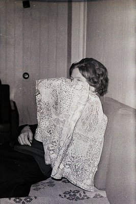 Vintage black-and-white portrait of a seated child hiding behind an ornate, floral-patterned shawl. Indoor setting with plain...