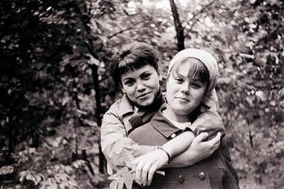 Two children pose closely in a wooded area, mid-20th century. Boy wears a short-sleeved jacket with a belt; girl has a headsc...