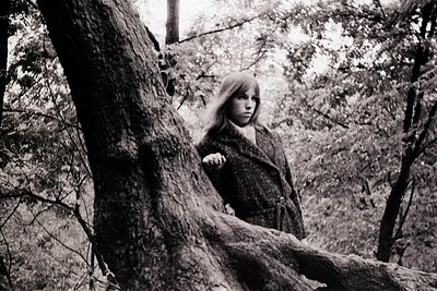 Young girl in textured coat leans against a tree trunk in a dense forest, black-and-white monochrome. Mid-20th century street...