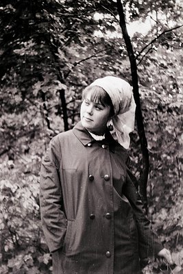 Young woman in a buttoned coat and headscarf stands outdoors among autumn foliage, mid-20th century. Her attire suggests a Eu...