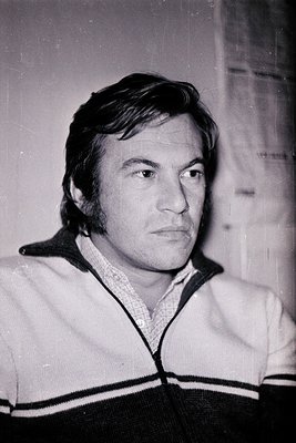 Portrait of a man in a 1970s-style sweater with a V-neck and horizontal stripes. Short, wavy hair and a neutral expression. I...