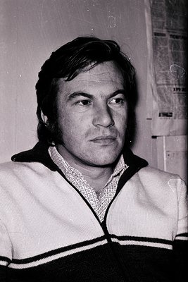 Portrait of a man in a 1970s-style sweater with black-and-white stripes, seated indoors with a newspaper background. His shor...