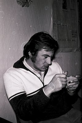 Mid-century man in a sweater vest and watch, examining a small handheld device—likely a camera or calculator—against a plain ...