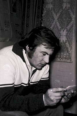 Man in 1970s-style track jacket examines a small object, possibly a camera or watch, indoors. Patterned wallpaper with geomet...