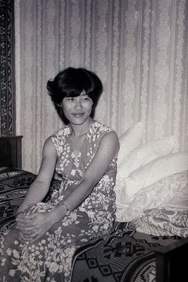Vintage black-and-white portrait of a woman in a floral-patterned dress, seated on a patterned bed with sheer curtains in the...