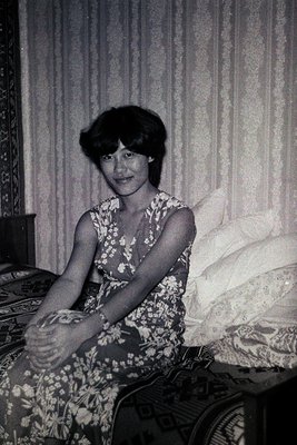 Vintage black-and-white portrait of a woman seated on a patterned bed, holding a small object in her hands. Floral dress with...