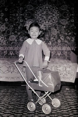 Mid-20th century black-and-white portrait of a young girl in a vintage dress, pushing a toy stroller with a fabric bag. Intri...